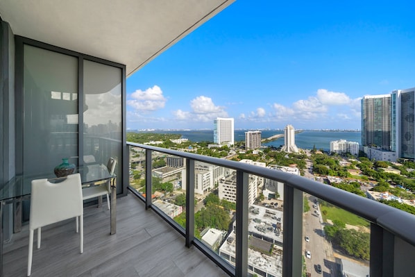 A balcony with a glass table and chair, offering a view of a cityscape with buildings and a body of water.