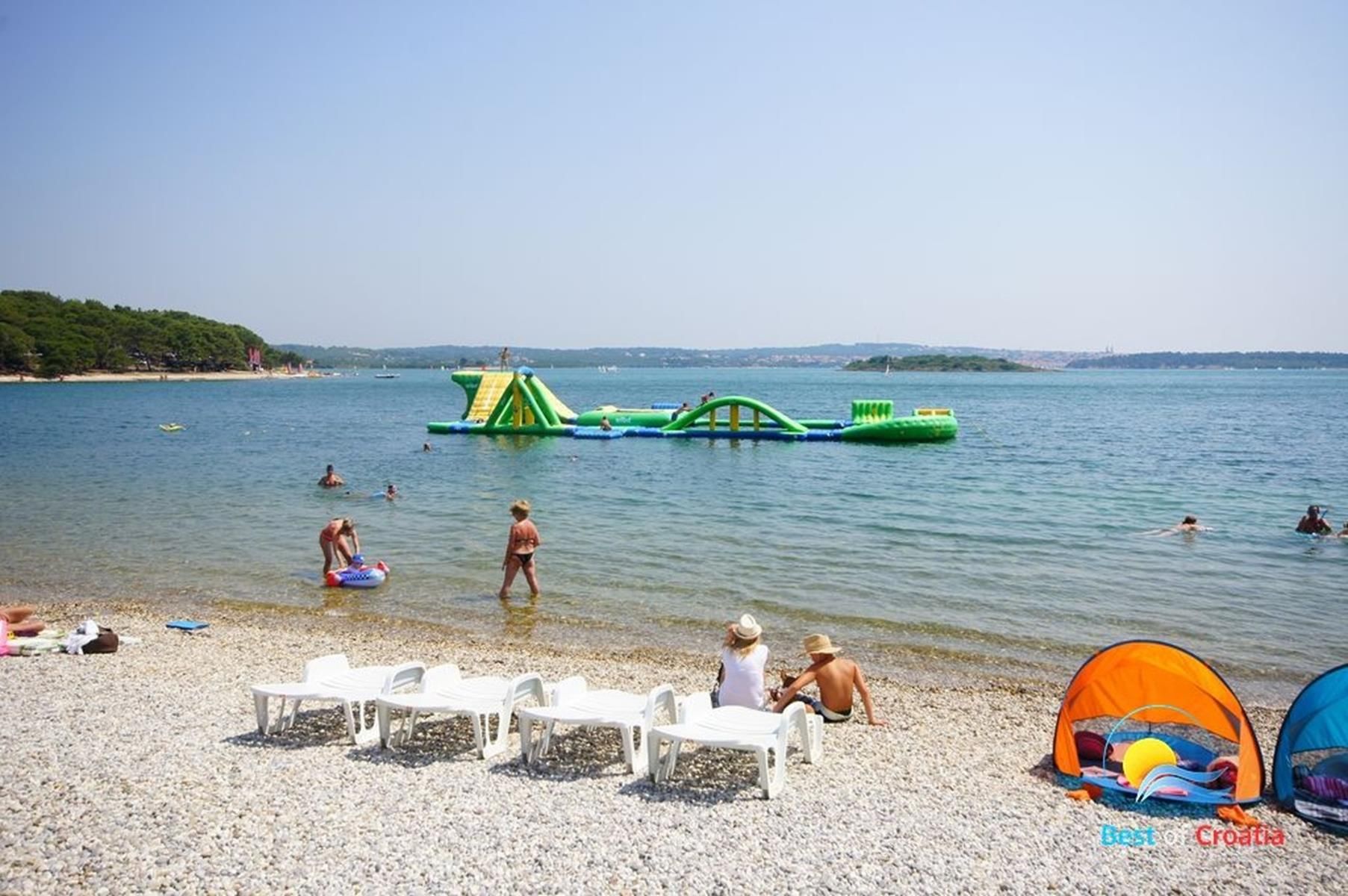 Una playa de guijarros con sillas blancas de plástico, una estructura hinchable verde para juegos y una carpa.
