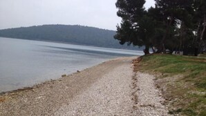 Ein Kiesweg führt zu einem See, begleitet von Bäumen auf der rechten Seite und einem bewaldeten Hügel im Hintergrund.