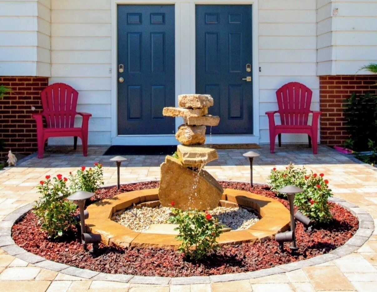 A stone water feature with a cascading design, surrounded by a circular stone border, with red chairs and a building with double doors in the background.