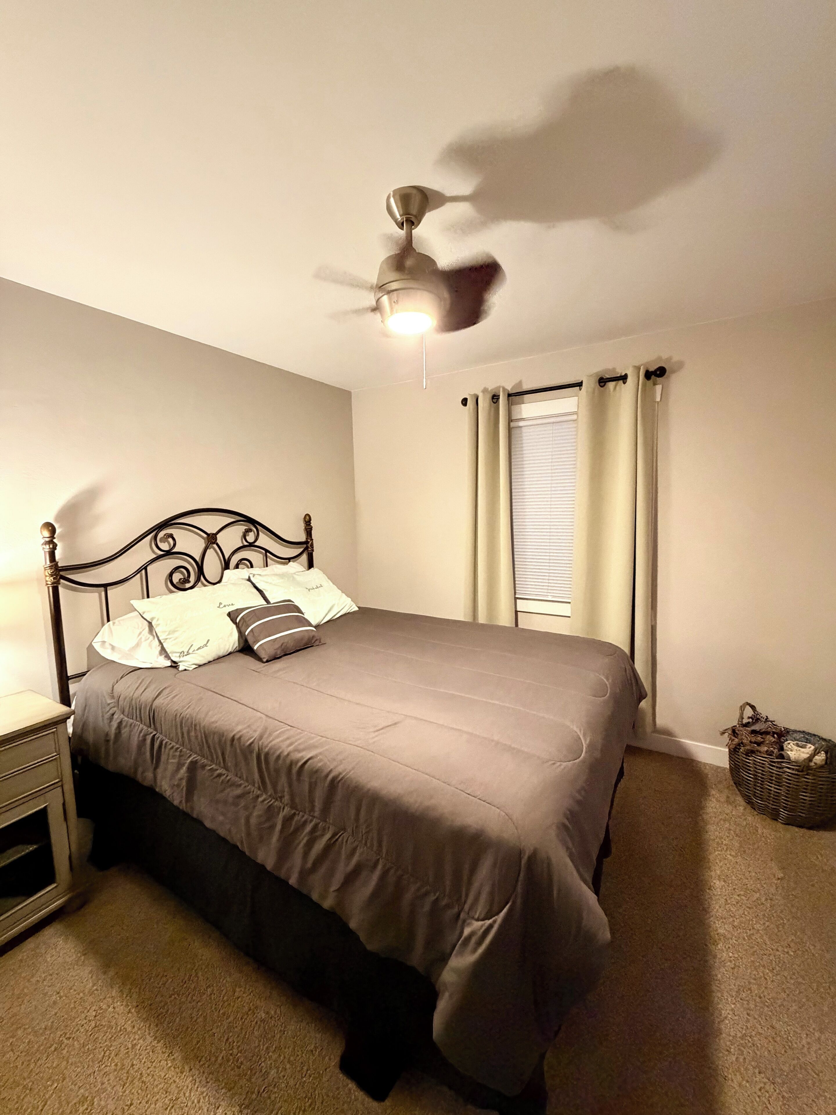 Un dormitorio con cama, mesita de noche, ventilador de techo y ventana con cortinas.