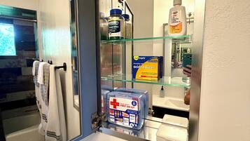 Een badkamer met een glazen plankje waarop toiletartikelen, een verbanddoos en een fles handdesinfectiemiddel staan.
