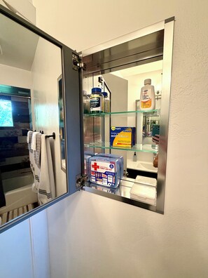Een badkamer met een glazen plankje waarop toiletartikelen, een verbanddoos en een fles handdesinfectiemiddel staan.