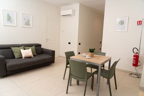 A room with a grey sofa, a wooden table with chairs, a small potted plant, and a fire extinguisher mounted on the wall. - Via Niccolò Tommaseo 4 - appartamento 1 (Napoli)
