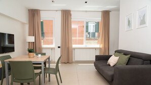 Ett modernt vardagsrum med en soffa, ett matbord med stolar, en platt-TV som är monterad på väggen och stora fönster med gardiner.