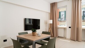 Ett modernt hotellrum med ett matbord i trä, gröna stolar, en platt-TV, en krukväxt, en lampa och ett fönster med gardiner.