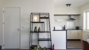 A modern kitchen with white cabinets, a black metal shelf unit, and a small dining area with a chair. - Seapoint, apartment with balcony and sea view (Zandvoort)
