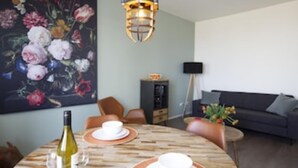 A dining area with a wooden table set for two, a bottle of wine, and a large floral painting on the wall. - Seapoint, apartment with balcony and sea view (Zandvoort)