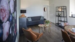 A modern living room with a grey sofa, a brown leather chair, a small round table with a vase, and a dining area with a table and chairs. - Seapoint, apartment with balcony and sea view (Zandvoort)
