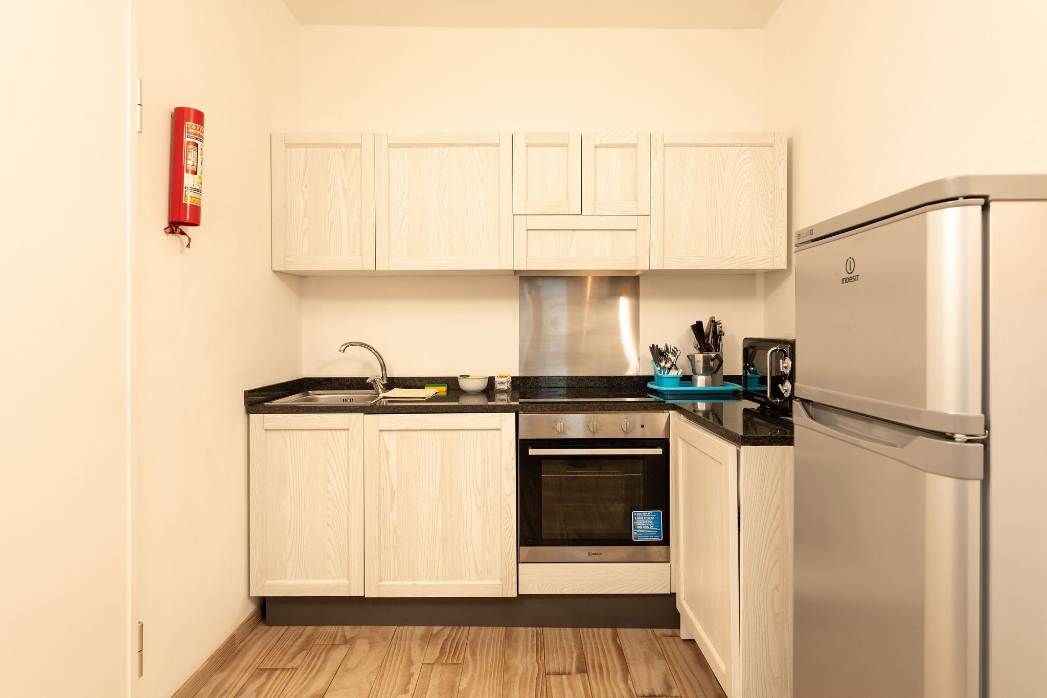 A compact kitchen with light wooden cabinets, a stainless steel oven, a refrigerator, and a fire extinguisher mounted on the wall.