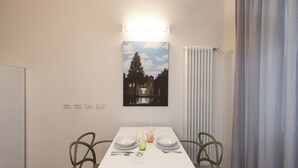 Un coin repas avec une table blanche, deux chaises et un tableau représentant une maison et un arbre accroché au mur.