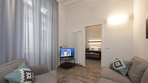 Un salon moderne doté d’un téléviseur à écran plat, de deux canapés ornés de coussins à motifs, d’un plancher en bois et d’un lit visible au fond.