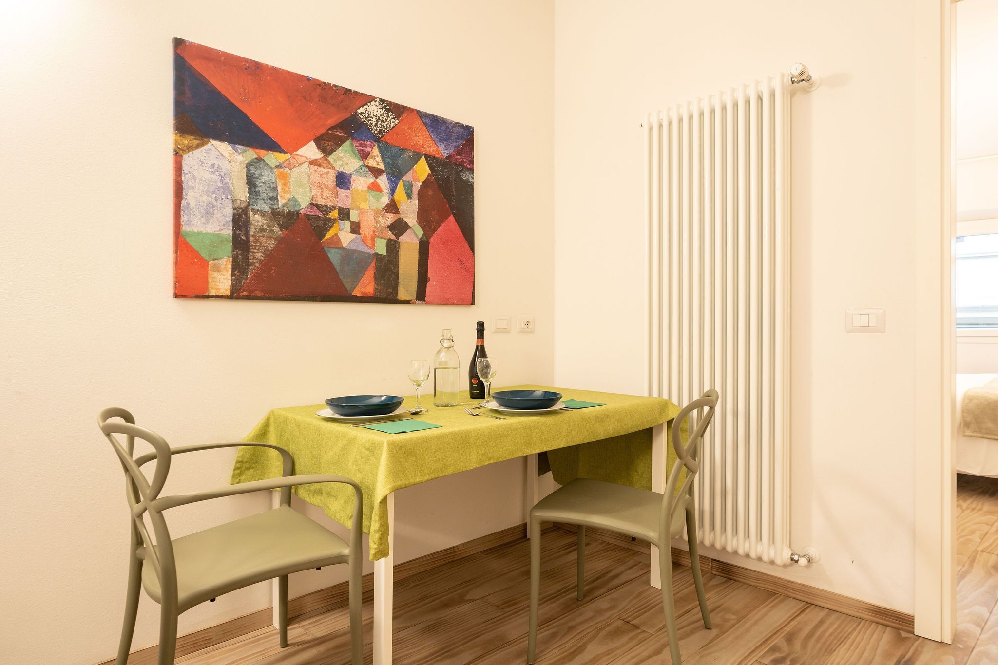 A dining area with a table set for two, a colorful abstract painting on the wall, and a radiator.