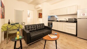 A compact living space with a black leather sofa, a small round table, a dining area with a green tablecloth, and a kitchenette with white cabinets and appliances.