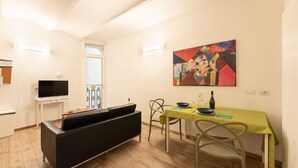 A modern living room with a black sofa, a dining table with a green tablecloth, and a colorful abstract painting on the wall.