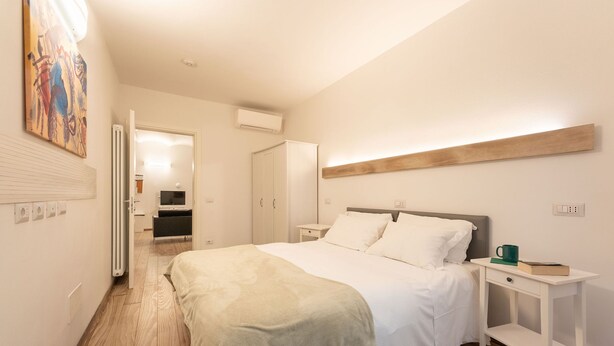 A bedroom with a bed, bedside tables, a nightstand, a radiator, and a door leading to another room.