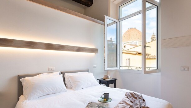 A neatly made bed with white linens, a bedside table with a hat, and a window with a view of a dome and buildings.