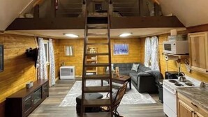 A loft-style room with a wooden floor, a loft bed, a kitchen with a stove and microwave, and a seating area with a couch and a coffee table. - Rustic Cabin at 17.5 Chena Hot Springs Rd. (Fairbanks)