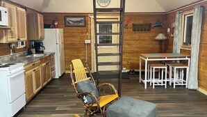 A compact kitchen with wooden cabinets, a stove, and a refrigerator. There's a dining area with a table and chairs, and a rocking chair. - Rustic Cabin at 17.5 Chena Hot Springs Rd. (Fairbanks)