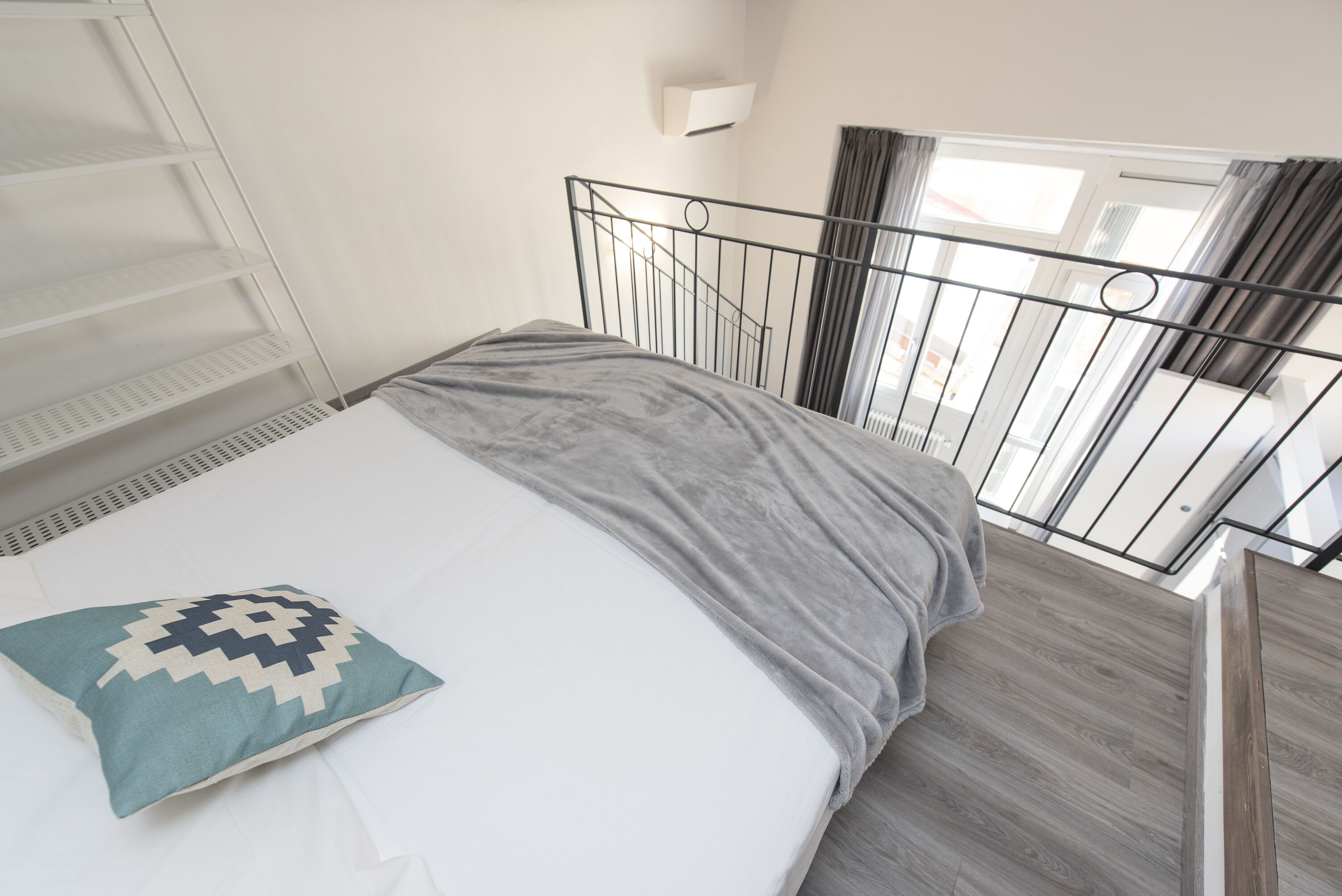 A bedroom with a bed, a grey blanket, a white pillow, a metal headboard, a window with curtains, and a white shelf unit.