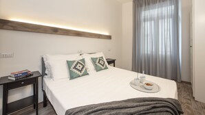 A neatly made bed with white bedding, two pillows with geometric patterns, a bedside table with a book, a cup, and a bowl of cereal.