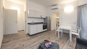 Cocina compacta con armarios blancos, horno integrado y refrigerador. Cuenta con una mesa de comedor con cuatro sillas y una pequeña zona de estar con un banco.