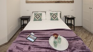Un dormitorio con cama, mesitas de noche, una mesita auxiliar, una colcha morada, una bandeja con tazas y un bol, y una lámpara de pared.