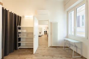 A hallway with a wooden floor, a white radiator, a window with blinds, and a clear desk with a chair.
