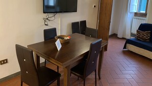 A dining area with a wooden table, six chairs, a television mounted on the wall, and a window with a view of the outside.