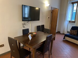 A dining area with a wooden table, six chairs, a television mounted on the wall, and a window with a view of the outside.