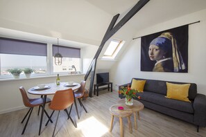 Un salon moderne comprenant un coin repas, un canapé gris agrémenté de coussins jaunes, un tableau représentant une femme et une lucarne.