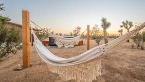 A hammock in a desert setting with wooden posts, surrounded by sparse vegetation and a building in the background.