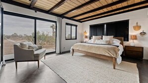 A spacious bedroom with a large bed, a sitting area with a chair, and a view of the desert landscape through large windows.