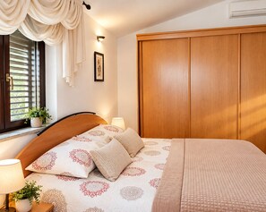 A bedroom with a bed, bedside lamps, a window with curtains, a wooden wardrobe, and a framed picture on the wall.