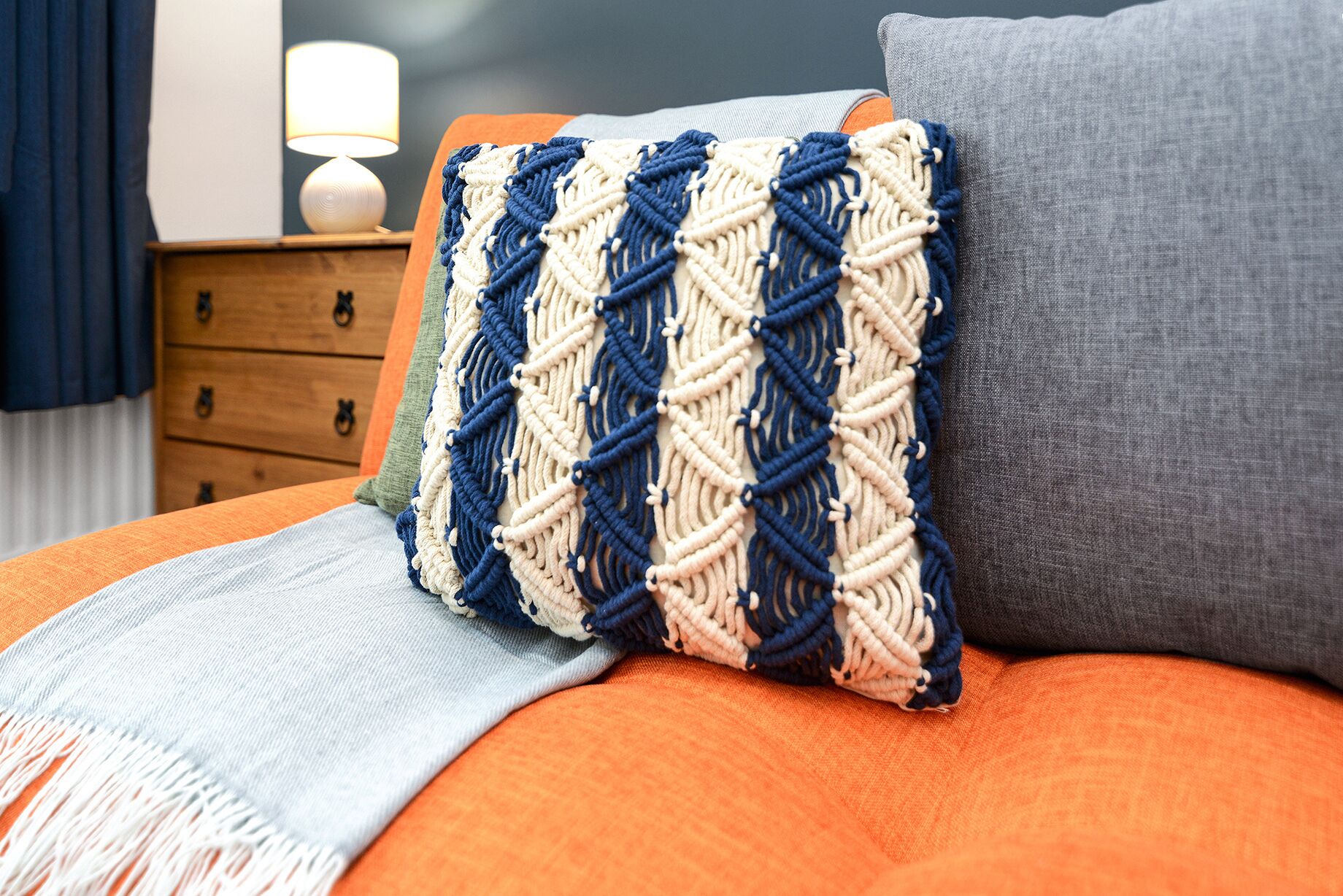 A bed with an orange blanket, a grey pillow, and a macrame pillow.