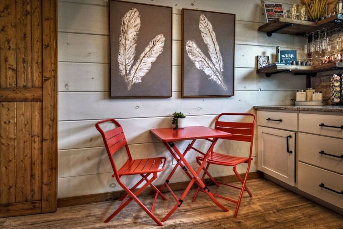 Een gezellige keuken met houten kastjes, een rode metalen tafel en twee rode stoelen. Twee lijstwerken met veerverbeelding hangen aan de muur.