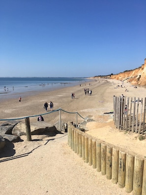 En sandstrand med folk som promenerar och solar, träkonstruktioner och en klarblå himmel.