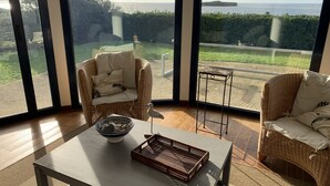 A room with large windows offering a view of a grassy area and the sea. There is a wicker chair with cushions, a small table with a bowl and a tray, and a metal stool.