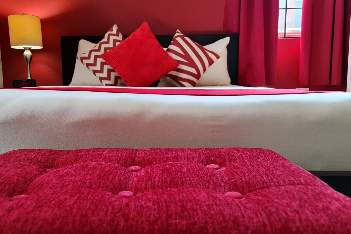 A hotel room with a bed, pillows, a lamp, and a red ottoman.
