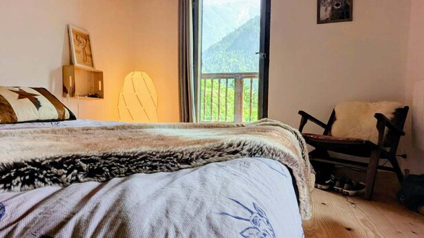 Un dormitorio con cama, una silla, techo de madera, ventana con vista a la montaña y una puerta.