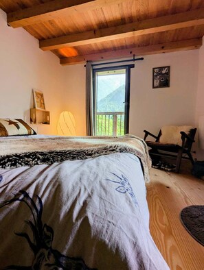 Ein Schlafzimmer mit Bett, Sessel, Holzboden und einem Fenster mit Blick auf die Berge sowie einer Tür.