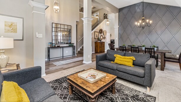 Amplia sala de estar con un sofá gris, una mesa de centro de madera y un tapete estampado. Una escalera conduce a un comedor con mesa y sillas de madera.