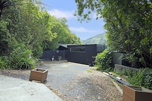 Une allée de gravier mène à un bâtiment en bois sombre, doté d’une fenêtre, et qui est entouré d’une végétation luxuriante, avec une montagne en arrière-plan.