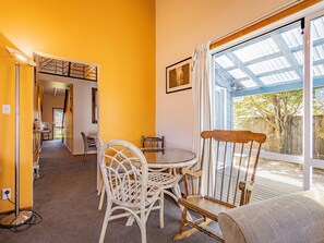 A room with a round glass table, wicker chairs, a wooden rocking chair, and a large window with a view of a wooden deck and trees.
