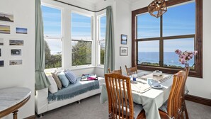 A dining area with a view of the sea, a bay window, and a seating area with cushions and a rug. - The Professor's Beach House - Brighton Home (Dunedin)