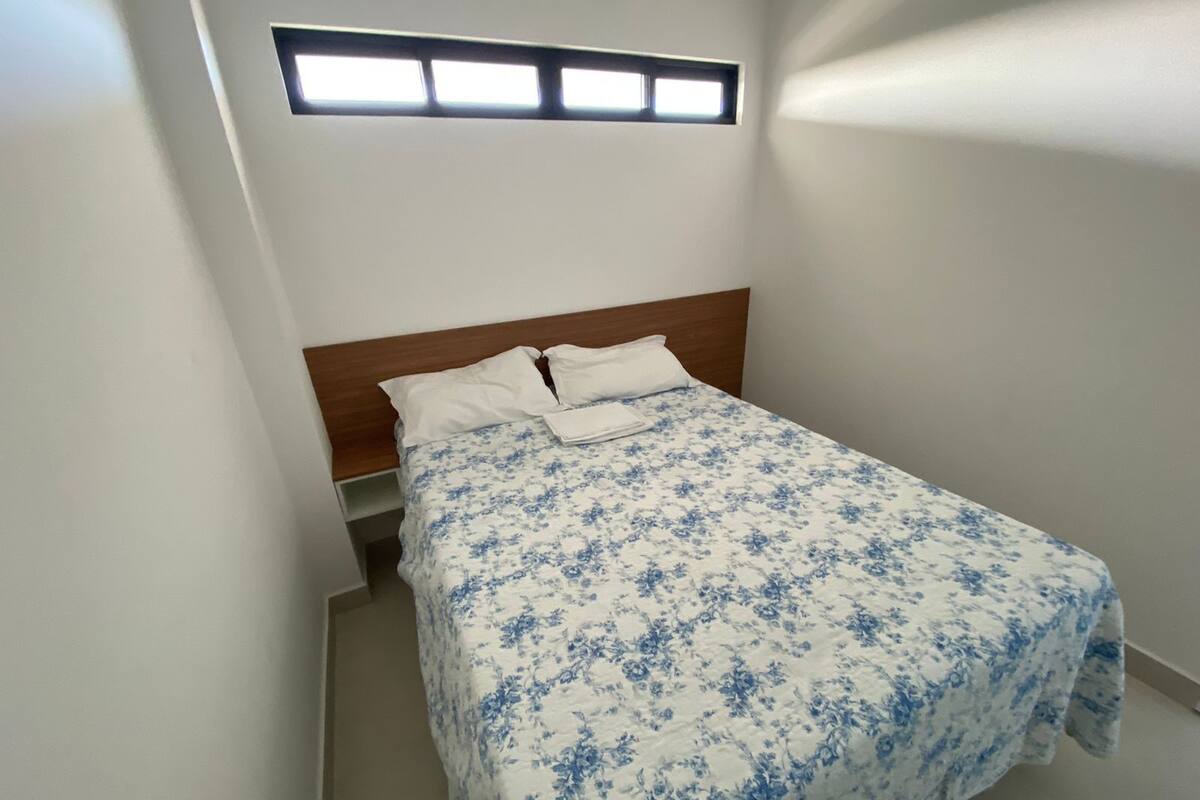 A bedroom with a bed, a wooden headboard, a window with blinds, and a small bedside table.