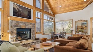 A cozy living room with a stone fireplace, wooden beams, and large windows offering a mountain view. - Fairway Vista: Massive Mountain Views w/ hot tub! (Mountain Village)