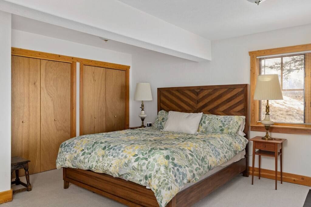 A bedroom with a wooden bed, a nightstand, a lamp, and a window with blinds.