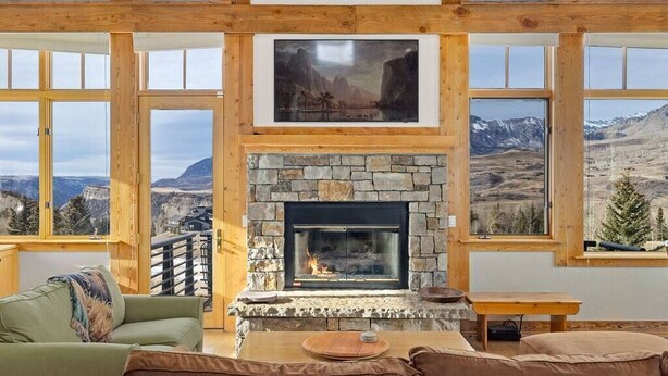 A cozy living room with a stone fireplace, wooden beams, and large windows offering mountain views. - Fairway Vista: Massive Mountain Views w/ hot tub! (Mountain Village)