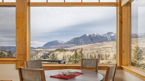 A dining area with a round table and four chairs, a bowl with a red placemat, and a view of mountains through large windows. - Fairway Vista: Massive Mountain Views w/ hot tub! (Mountain Village)